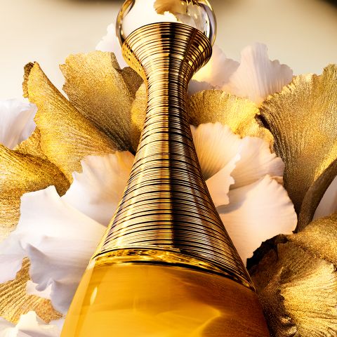 Eine Nahaufnahme eines goldenen Parfümflakons mit strukturiertem, spiralförmigem Design vor zartweißen und goldenen Blütenblättern im Hintergrund. Das Bild strahlt Luxus und Eleganz aus.