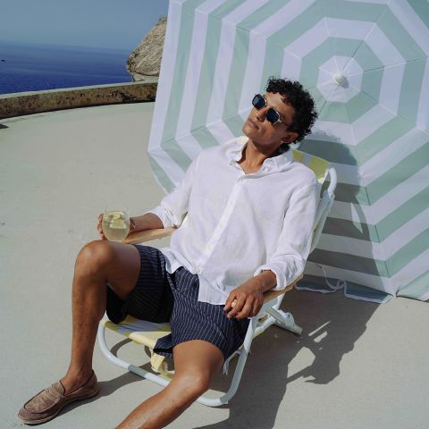 Ein Mann mit Sonnenbrille und weißem Hemd entspannt sich an einem sonnigen Tag am Meer auf einem Liegestuhl mit einem Drink unter einem gestreiften Sonnenschirm.