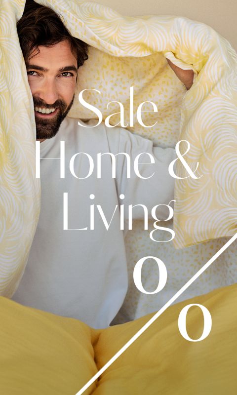 Ein lächelnder Mann lugt unter einer gelb gemusterten Decke hervor. Der weiße Text über dem Bild lautet: "Sale Home & Living" mit einem großen Prozentzeichen darunter.