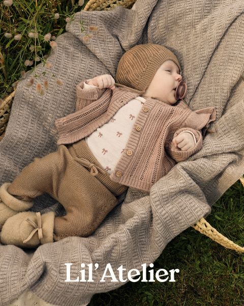 Ein Baby, das eine braune Strickmütze, eine Strickjacke und eine Hose trägt, liegt auf einer strukturierten beigen Decke in einem Korb im Freien. Das Baby hat einen Schnuller und Füßchen. Der Text am unteren Rand lautet "Lil' Atelier.