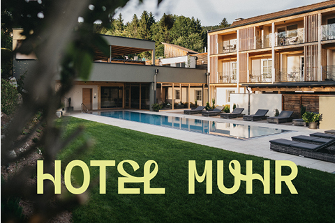 Ein modernes Hotel mit Holzbalkonen blickt auf einen von Liegestühlen und grünem Gras umgebenen Pool, über dem in großen, hellgelben Buchstaben "HOTEL MUHR" steht.