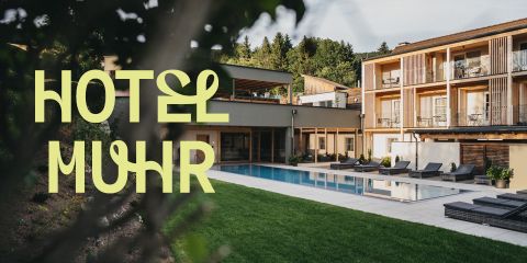 Außenansicht des Hotels Muhr mit einem modernen Gebäude, Balkonen, Liegestühlen am Pool, grünem Rasen und Bäumen im Hintergrund. Großer stilisierter gelber Text lautet "HOTEL MUHR.