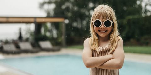 Lächelndes Kind mit blondem Haar und runder Sonnenbrille steht mit verschränkten Armen vor einem Swimmingpool an einem regnerischen Tag, mit Liegestühlen und Bäumen im Hintergrund.