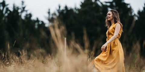 Eine Frau in einem gelben Kleid lächelt, während sie durch ein grasbewachsenes Feld mit hohen Bäumen im Hintergrund geht.