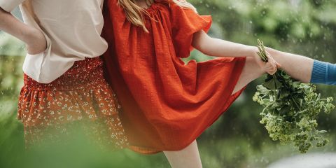 Zwei Personen stehen im Freien dicht beieinander, eine trägt ein rotes Kleid, die andere ein weißes Hemd und einen gemusterten Rock. Eine dritte Hand hält grünes Laub in der Nähe des Beins der Person im roten Kleid. Im Hintergrund ist Grünzeug zu sehen.