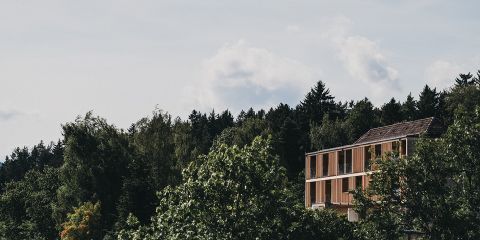 Ein modernes Gebäude mit Holzbalkonen, die teilweise von hohen grünen Bäumen verdeckt werden, vor dem Hintergrund eines dichten Waldes und eines bewölkten Himmels.