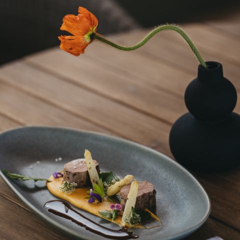 Ein modernes Gericht mit zwei Fleischscheiben, weißem Spargel, essbaren Blumen und Soßen wird kunstvoll auf einem grauen ovalen Teller neben einer schwarzen Vase mit einer einzelnen orangefarbenen Blume auf einem Holztisch angerichtet.