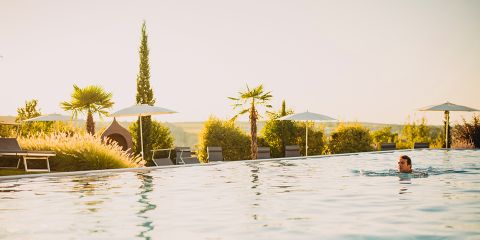 Ein Mann schwimmt in einem Außenpool, umgeben von Liegestühlen, weißen Sonnenschirmen und Palmen, mit einer malerischen Landschaft und goldenem Sonnenlicht im Hintergrund.
