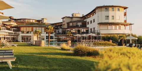 Ein modernes Resort-Hotel mit mehreren Stockwerken und Balkonen überblickt eine gepflegte Rasenfläche mit Liegestühlen, Palmen, einem Swimmingpool und schattigen Terrassenbereichen unter einem klaren Himmel.
