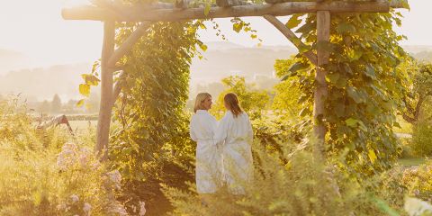 Zwei Frauen in weißen Gewändern stehen unter einer hölzernen, mit grünen Ranken bewachsenen Laube, umgeben von üppigen Pflanzen und Blumen, und genießen eine sonnige Gartenszene.