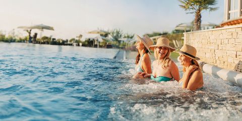 Vier Frauen in Badeanzügen und Sonnenhüten sitzen am Rande eines Pools und unterhalten sich, lächeln und genießen einen sonnigen Tag. Im Hintergrund sind Palmen und Sonnenschirme zu sehen.