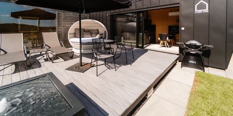 Eine moderne Außenterrasse mit Liegestühlen, einer runden Liege, einem Esstisch mit Stühlen, einem Grill und einem großen Sonnenschirm. Glasschiebetüren führen in das stilvolle Innere der Hütte. Gras und Trittsteine begrenzen die Terrasse.