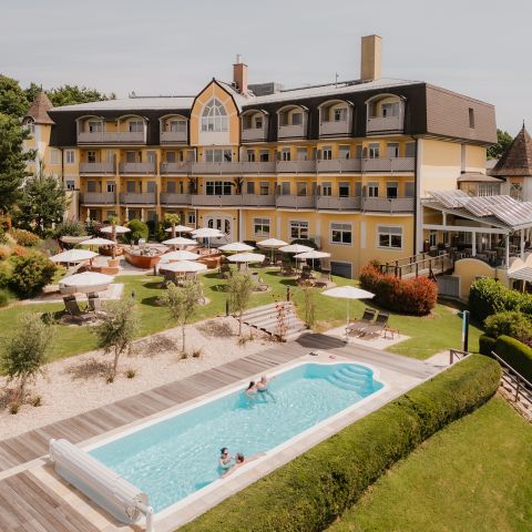 Ein gelbes Hotel mit Balkonen blickt auf einen angelegten Garten mit Liegestühlen, Sonnenschirmen und einem langen Außenpool. Mehrere Personen entspannen sich am Pool und auf der Liegewiese unter sonnigem Himmel.