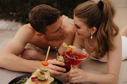Ein Mann und eine Frau liegen im Freien eng beieinander, lächeln und stellen Blickkontakt her, während sie bunte Cocktails halten. Im Vordergrund ist das Essen zu sehen, das eine entspannte, romantische Atmosphäre schafft.