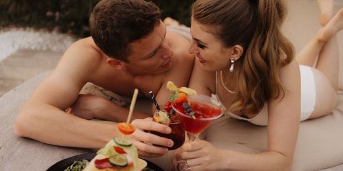 Ein Mann und eine Frau in Badeanzügen liegen eng beieinander im Freien, lächeln und stellen Blickkontakt her, während sie mit bunten Cocktails anstoßen. Vor ihnen steht ein Teller mit Appetithäppchen mit Obst und Gemüse.