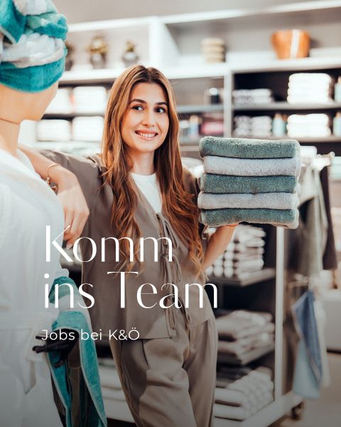 Eine lächelnde Frau in einem Geschäft hält einen Stapel gefalteter Handtücher neben einer Schaufensterpuppe. Der Text auf dem Bild lautet: "Komm ins Team - Jobs bei K&Ö". Im Hintergrund befinden sich Regale mit weiteren Handtüchern.