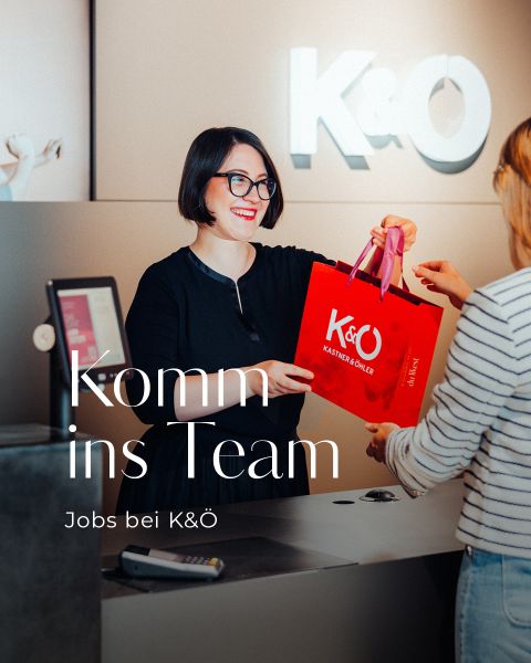 Ein lächelnder Mitarbeiter überreicht einem Kunden an einer modernen Theke eine rote K&Ö-Einkaufstasche. In großer Schrift steht "Komm ins Team" und "Jobs bei K&Ö". An der Wand im Hintergrund ist das K&Ö-Logo zu sehen.