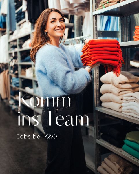 Eine lächelnde Frau in einem blauen Pullover stapelt rote Schals auf einem Regal in einem Bekleidungsgeschäft. Der Text auf dem Bild lautet: "Komm ins Team. Jobs bei K&Ö." Um sie herum sind weitere gefaltete Kleidungsstücke zu sehen.