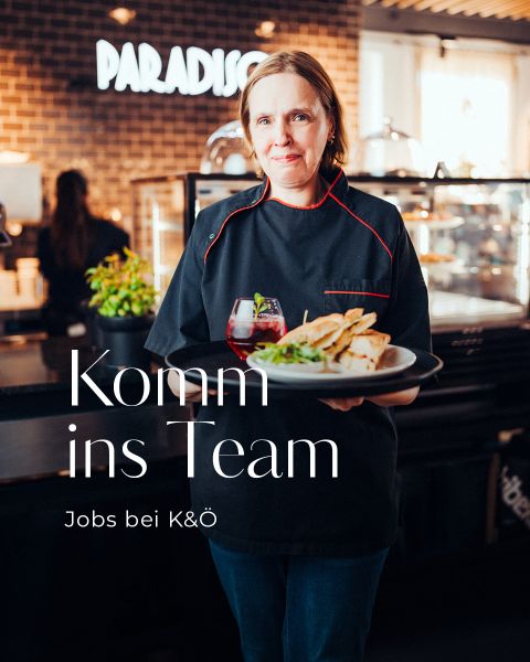 Eine Frau in schwarzer Kochuniform steht in einem Restaurant und hält ein Tablett mit einem Getränk und einem Teller mit Essen. Der Text auf Deutsch lautet: "Komm ins Team. Jobs bei K&Ö." Backsteinmauer und "PARADISO"-Schild im Hintergrund.