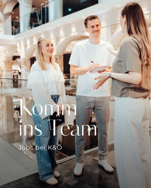Zwei junge Erwachsene lächeln und unterhalten sich mit einer Frau in einem modernen, eleganten Innenbereich. Der Text auf Deutsch lautet "Komm ins Team. Jobs bei K&Ö." lädt dazu ein, das Team von K&Ö zu verstärken.