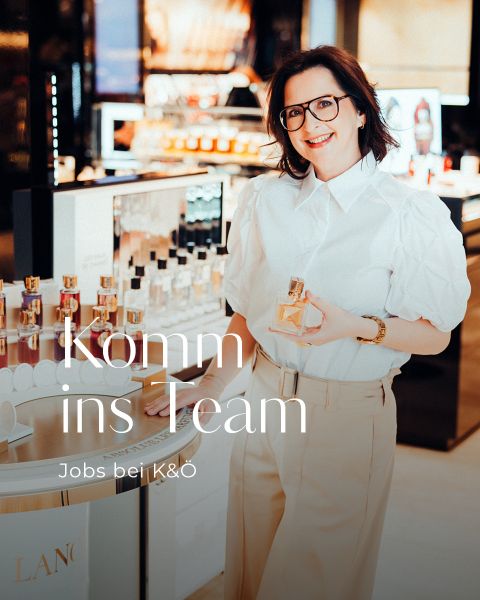 Eine lächelnde Frau in weißer Bluse und beiger Hose steht in einem Parfümgeschäft und hält eine Parfümflasche in der Hand. Der Text auf dem Bild lautet: "Komm ins Team - Jobs bei K&Ö.