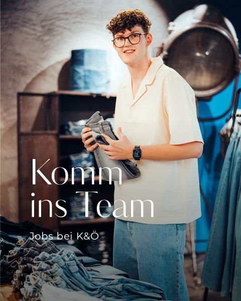Eine junge Person mit lockigem Haar und Brille lächelt, während sie in einem Bekleidungsgeschäft Jeans faltet. Der Text lautet: "Komm ins Team. Jobs bei K&Ö." Im Hintergrund sind Jeansartikel und Ladenregale zu sehen.