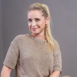 Eine Frau mit langen blonden Haaren zu einem Pferdeschwanz, einem beigefarbenen Strickoberteil und Goldschmuck sitzt lächelnd vor einem schlichten grauen Hintergrund.