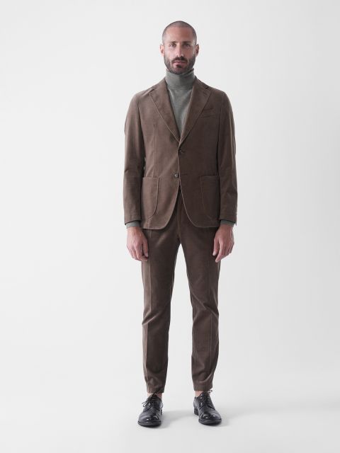 Ein Mann steht mit dem Gesicht nach vorne, trägt einen braunen Anzug mit Blazer und passender Hose, einen grauen Rollkragenpullover und schwarze Anzugschuhe vor einem schlichten weißen Hintergrund.