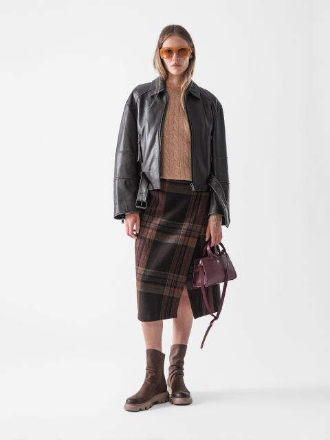 Eine Frau, die eine schwarze Lederjacke, einen beigen Pullover, einen karierten Midirock, braune Stiefel und eine orangefarbene Sonnenbrille trägt, steht vor einem weißen Hintergrund und hält eine kleine weinrote Handtasche.