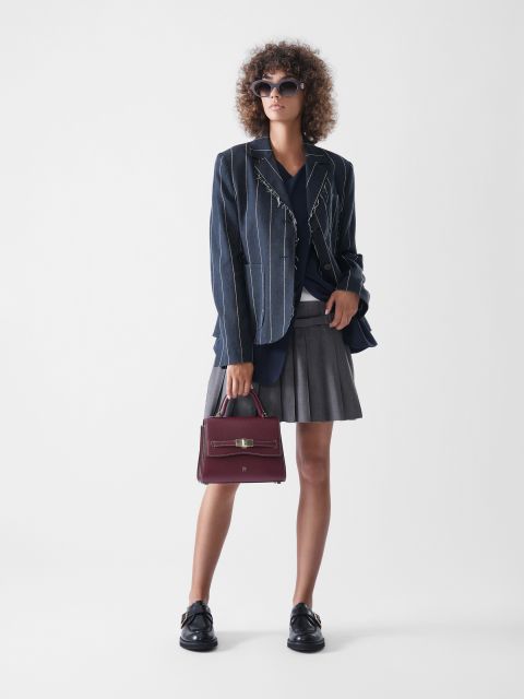 Eine Frau mit lockigem Haar trägt eine Sonnenbrille, einen marineblauen Nadelstreifenblazer, ein marineblaues Oberteil, einen grauen Faltenrock und schwarze Schuhe. Sie hält eine kastanienbraune Handtasche in der Hand und steht vor einem schlichten weißen Hintergrund.