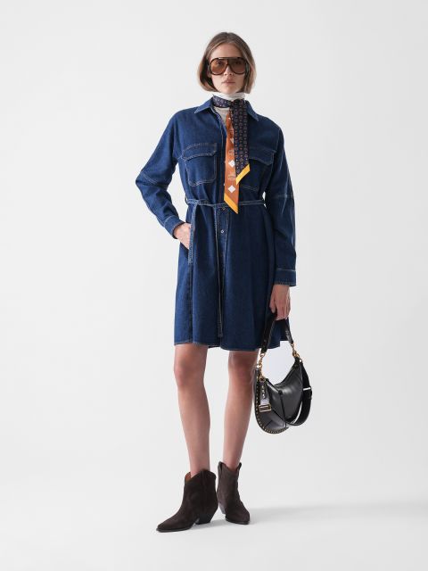 Eine Frau posiert vor einem schlichten Hintergrund und trägt ein Jeanskleid mit Gürtel, einen gemusterten Schal, eine große Sonnenbrille, braune Ankle Boots und eine schwarze Handtasche.