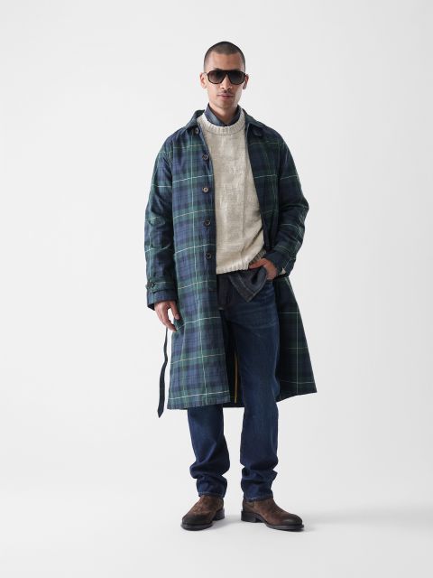 Ein Mann mit dunkler Sonnenbrille, einem langen grün-blau karierten Mantel, einem cremefarbenen Pullover, blauen Jeans und braunen Stiefeln steht selbstbewusst vor einem schlichten weißen Hintergrund.