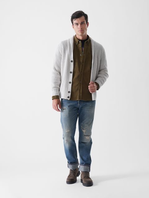 Ein Mann steht vor einem schlichten weißen Hintergrund und trägt eine cremefarbene Rippenstrickjacke über einem olivgrünen Hemd, blaue Jeans und braune Stiefel. Er blickt mit neutraler Miene nach vorne und hat eine Hand in der Tasche.