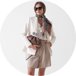 Eine Frau, die eine Sonnenbrille, einen gemusterten Schal, einen weißen Blazer, eine Rüschenbluse und beigefarbene Shorts trägt, hält eine braune Clutch-Bag mit einer silbernen Spange und posiert vor einem weißen Hintergrund.