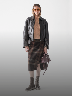 Eine Frau steht vor einem schlichten Hintergrund und trägt eine orangefarbene Sonnenbrille, eine schwarze Lederjacke, einen hellbraunen Zopfstrickpullover, einen braun karierten Rock, taupefarbene Stiefel und eine kleine braune Handtasche.