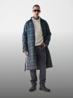 Ein Mann mit Sonnenbrille, einem langen grün-blau karierten Mantel über einem cremefarbenen Pullover, dunklen Jeans und braunen Stiefeln steht vor einem schlichten, hellgrauen Hintergrund.