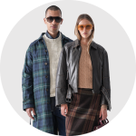 Ein Mann in einem grün karierten Mantel und einer Sonnenbrille steht neben einer Frau in einer schwarzen Lederjacke, einem hellbraunen Pullover, einem karierten Rock und einer bernsteinfarbenen Sonnenbrille. Beide blicken vor einem hellgrauen Hintergrund nach vorne.