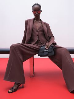 Ein Model in einem braunen Oversized-Anzug, einer Hose mit weitem Bein und schwarzen Absätzen sitzt auf einer Bank vor einem roten Boden und einer weißen Wand, trägt eine Sonnenbrille und hält eine schwarze HUGO-Handtasche in der Hand.