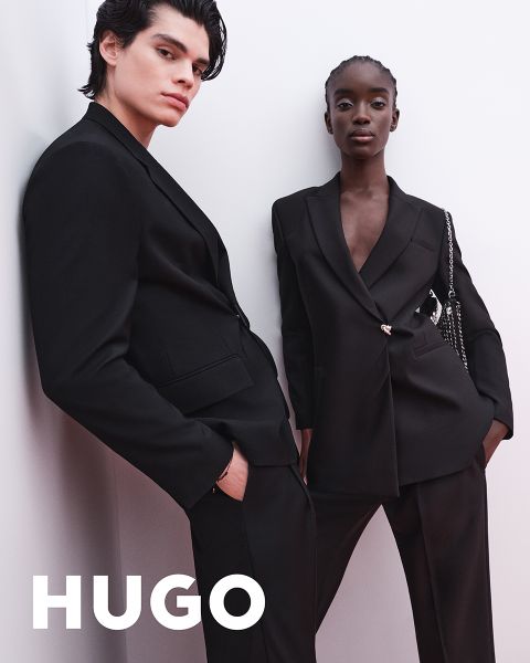 Zwei Models in schwarzen Anzügen posieren selbstbewusst vor einem schlichten hellen Hintergrund. Das HUGO Logo erscheint in fetten weißen Buchstaben in der unteren linken Ecke.