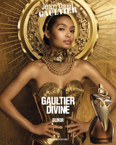 Eine Frau in einem goldenen Metallkleid und mit Schmuck posiert vor einem goldenen Sonnenschliff-Hintergrund mit einem Flakon des Parfums Jean Paul Gaultier Divine Elixir im Vordergrund. Der Name des Parfums ist in fetter weißer Schrift dargestellt.