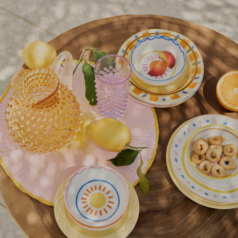 Ein runder Holztisch mit bunten Tellern, einem rosa Tischset, einem gemusterten gelben Glaskrug, einem rosa Glas, Zitronen, einer halbierten Orange und einem Teller mit kleinen runden Keksen. Das Sonnenlicht erzeugt weiche Schatten.