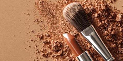 Ein Schminkpinsel und ein nudefarbener Lippenstift liegen auf einer mit losem braunem Puder bestreuten Fläche, die alle in ähnlichen Erdtönen gehalten sind.