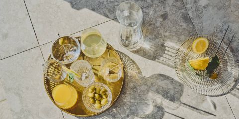 Ein goldenes Tablett mit verschiedenen Getränken, Oliven und einem Glas Orangensaft steht auf einer gefliesten Fläche im Sonnenlicht. In der Nähe steht ein Glasteller mit Zitronenscheiben und einem Blatt, daneben ein Glaskrug mit Wasser.