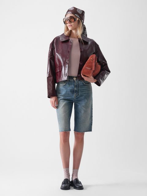 Eine Person, die eine kastanienbraune Lederjacke, ein beiges Oberteil, knielange Jeans-Shorts, schwarze Halbschuhe, eine Sonnenbrille und ein gemustertes Kopftuch trägt und eine braune Handtasche in der Hand hält, steht vor einem schlichten weißen Hintergrund.