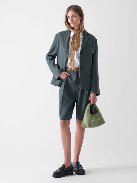 Eine Frau steht vor einem schlichten Hintergrund und trägt einen grünen Blazer und dazu passende knielange Shorts, einen beige gemusterten Schal, ein weißes Hemd, schwarze Halbschuhe und eine kleine grüne Handtasche.
