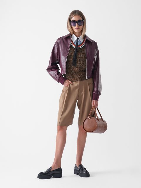 Eine Person mit Sonnenbrille trägt eine weinrote Lederjacke, eine braune Strickweste über einem Hemd mit Kragen und Krawatte, hellbraune knielange Shorts, schwarze Halbschuhe und eine runde braune Handtasche vor einem weißen Hintergrund.