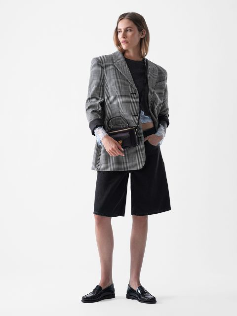 Eine Frau steht vor einem weißen Hintergrund und trägt einen grau karierten, übergroßen Blazer, ein schwarzes Top, lange schwarze Shorts, schwarze Halbschuhe und eine kleine schwarze Handtasche. Sie hat schulterlanges Haar und einen neutralen Gesichtsausdruck.