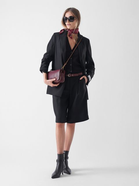Eine Frau steht vor einem hellen Hintergrund und trägt einen schwarzen Blazer, schwarze knielange Shorts, einen weinroten Schal, eine Sonnenbrille, Ankle Boots und eine Crossbody-Tasche.