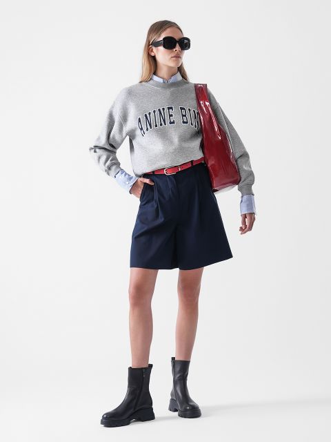 Eine Frau, die ein graues "NINE BIN"-Sweatshirt, blaue Shorts, schwarze Stiefel, eine Sonnenbrille und eine rote Umhängetasche trägt, steht selbstbewusst vor einem schlichten weißen Hintergrund.