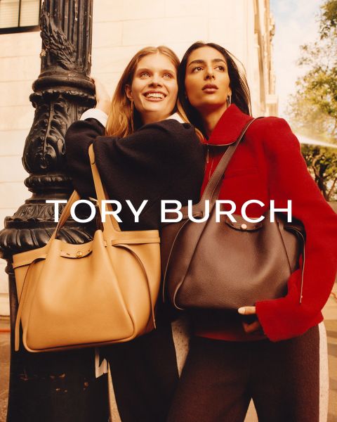 Zwei Frauen stehen Arm in Arm im Freien, lächeln, tragen elegante Mäntel und große Handtaschen von Tory Burch. Das Sonnenlicht hebt ihre Gesichter hervor. Der Text "TORY BURCH" erscheint über dem Bild.
